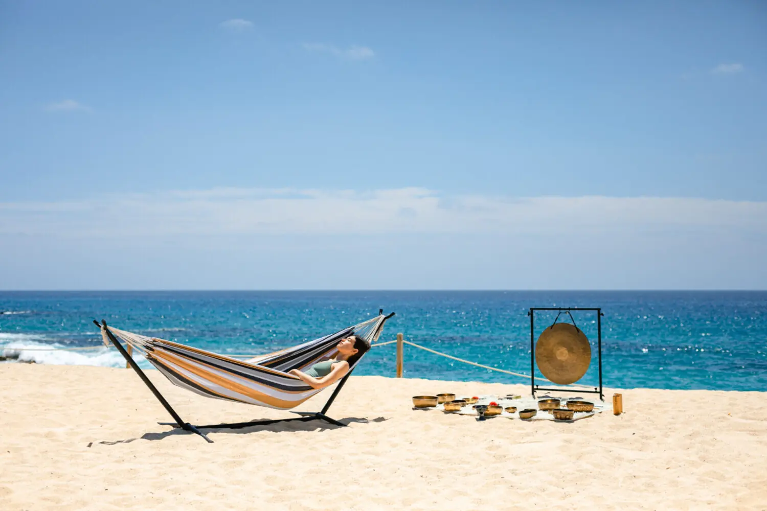 Person relaxes in striped hammock on sandy beach, with sound bowls and gong, facing ocean.