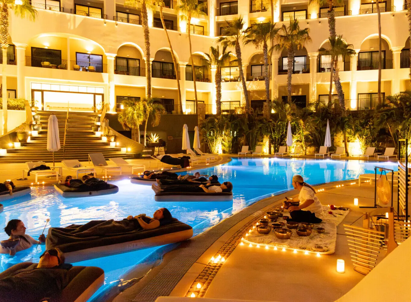 People lounge on floating beds in a lit resort pool at night, enjoying a relaxing sound bath experience.