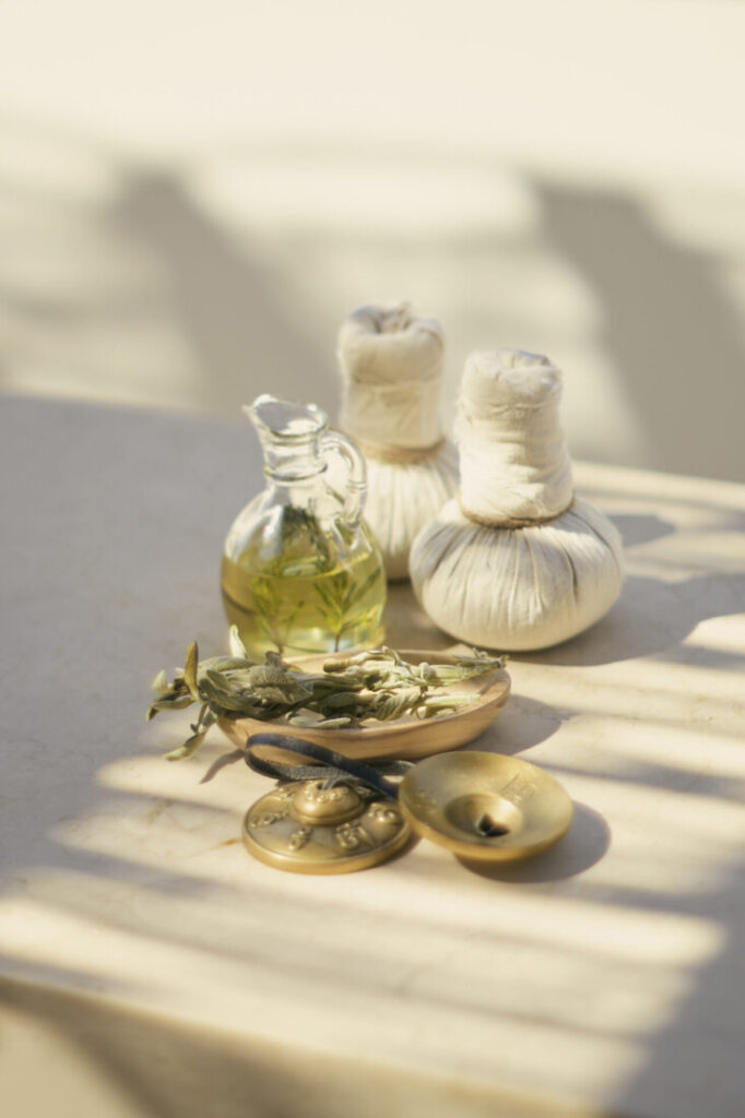Glass bottle of herbal oil with two wrapped massage compresses arranged on a sunlit spa table.