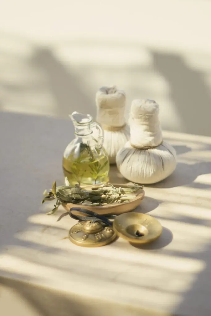 Glass bottle of herbal oil with two wrapped massage compresses arranged on a sunlit spa table.