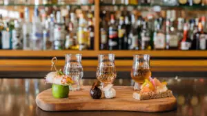 Three glasses of liquor paired with creative appetizers on wooden board, background of colorful bar shelves.