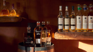 Botellas de licor surtidas exhibidas en barriles y estantes de madera, barra cálidamente iluminada con reflejos ámbar y dorados.