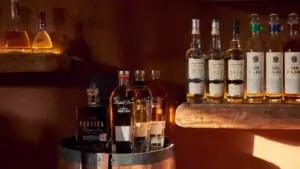 Assorted liquor bottles displayed on barrel and wooden shelves, warmly lit bar with amber and gold highlights.