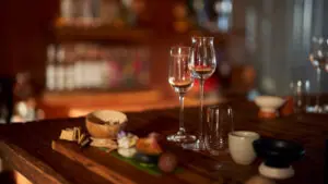 Two wine glasses, snacks, and ceramic cups arranged on a wooden bar counter in soft focus lighting.