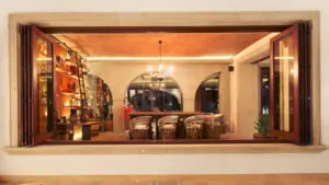 Warmly lit restaurant interior viewed through large open window, featuring wooden shelves, elegant chandeliers, and neatly set tables.