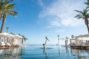 Parent tossing a child into an infinity pool at a palm-lined beachfront resort.