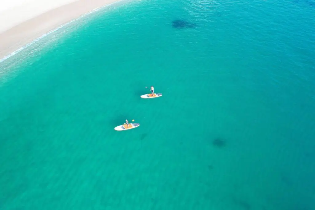 Aerial view of two people paddleboarding on clear turquoise water near a sandy shoreline.