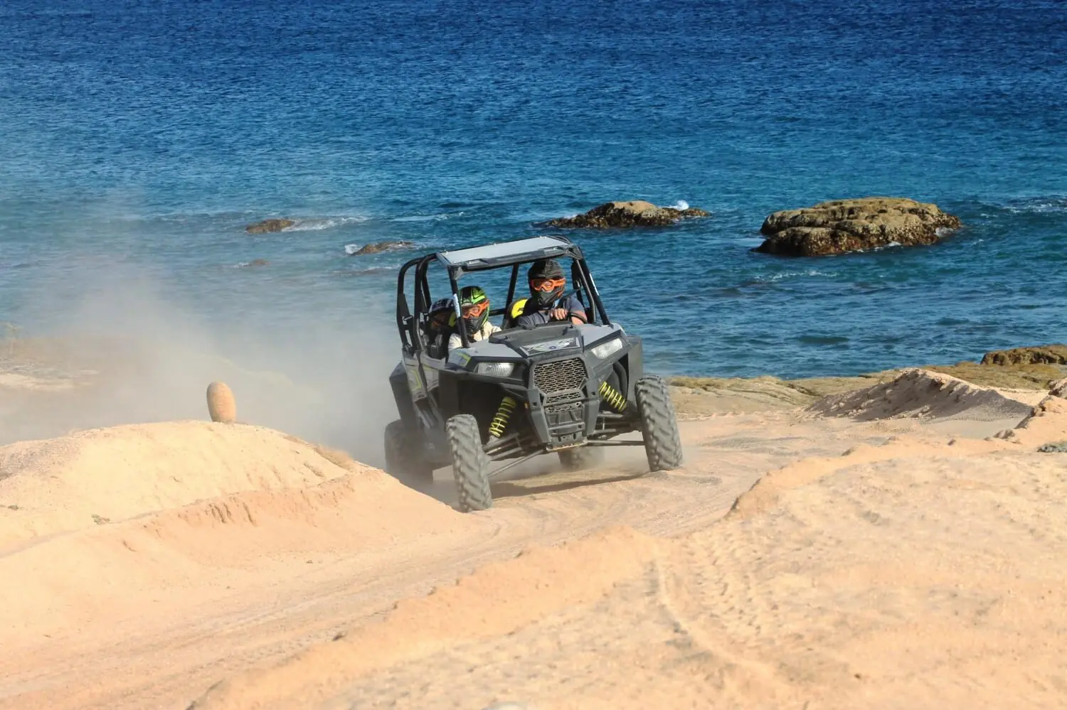 Couple drives off-road buggy along sandy coastline, kicking up dust with ocean waves nearby.