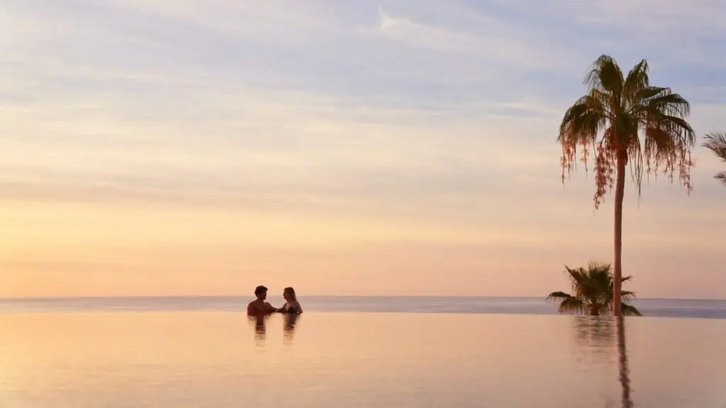Couple standing in an infinity pool at sunset, with palm trees and the ocean blending into the horizon.