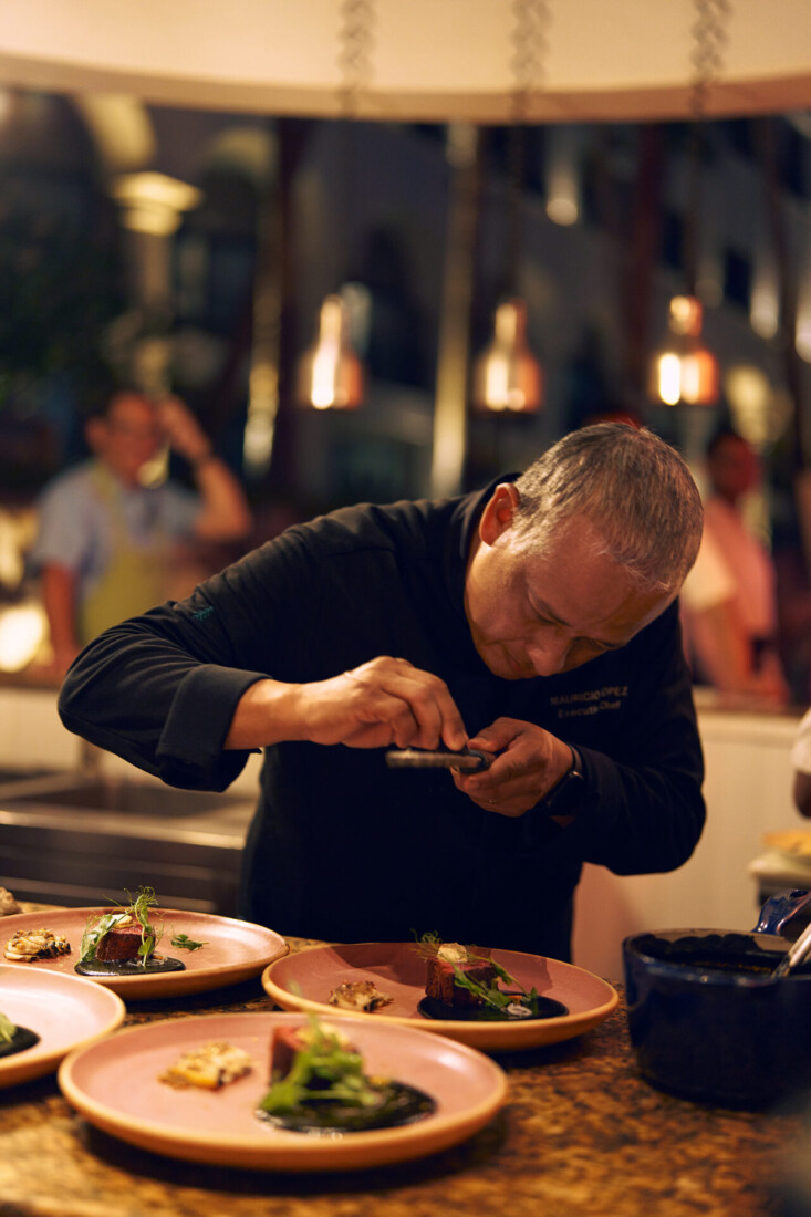 Chef in black coat prepares gourmet dishes on pink plates in warmly lit open kitchen.