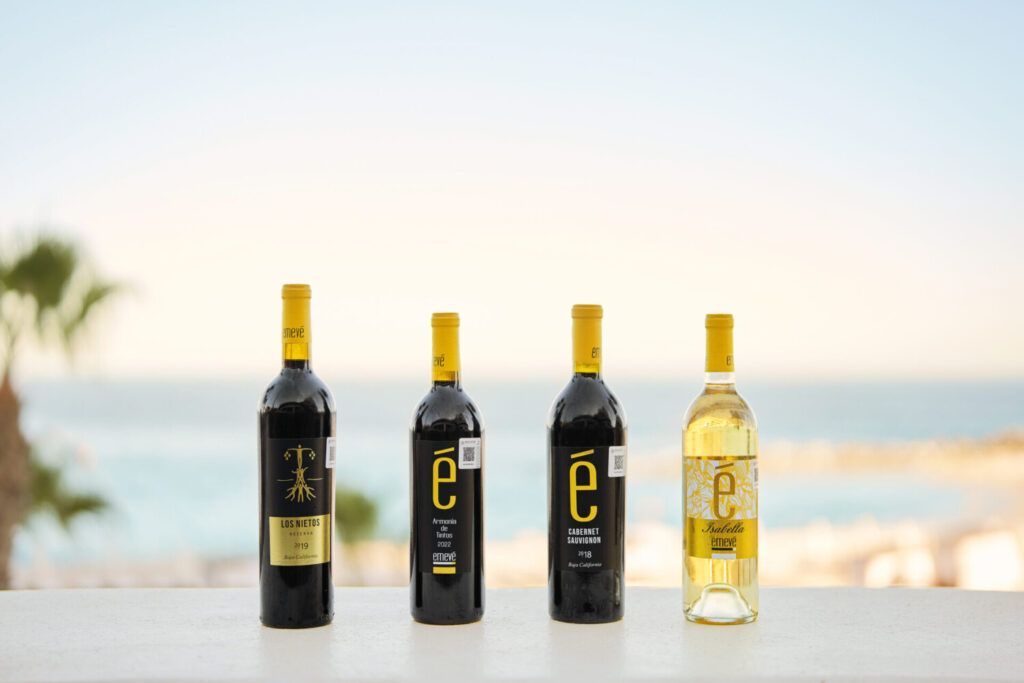 Four wine bottles displayed on a ledge with ocean views in the background, including red and white varieties.