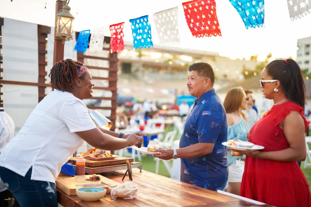 Outdoor food station where guests receive plates at a festive event with colorful banners and string lights.