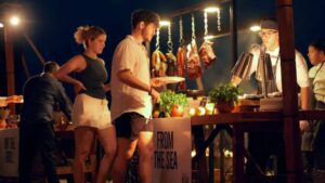 Los invitados piden comida en un mercado nocturno al aire libre con chefs asando y sirviendo platos bajo luces festivas.