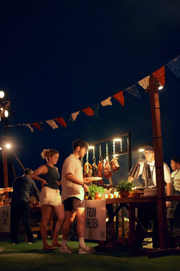 Guests ordering food at an outdoor night market with chefs grilling and serving dishes under festive lights.