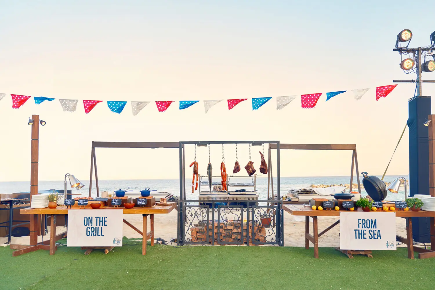 Beachfront grill station with seafood and meats, festive banners, and ocean view in the background.