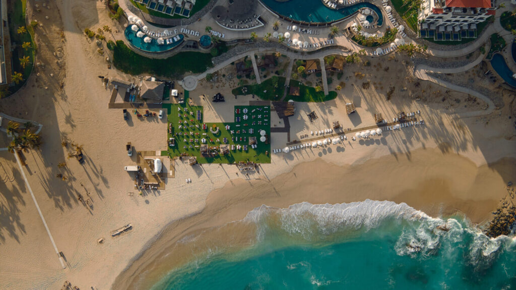 Aerial view of a beachfront resort with event setup on the sand, pools, lounge chairs, and turquoise ocean.