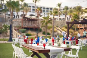 Configuración de evento al aire libre en un resort con largas mesas de comedor, vajilla roja, blanca y azul, palmeras y luces de cadena.