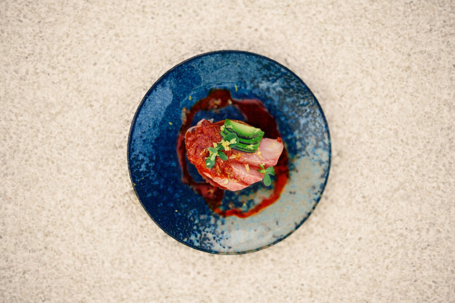 Top-down view of a gourmet plated dish with tuna, sauce, and microgreens served on a blue ceramic plate.