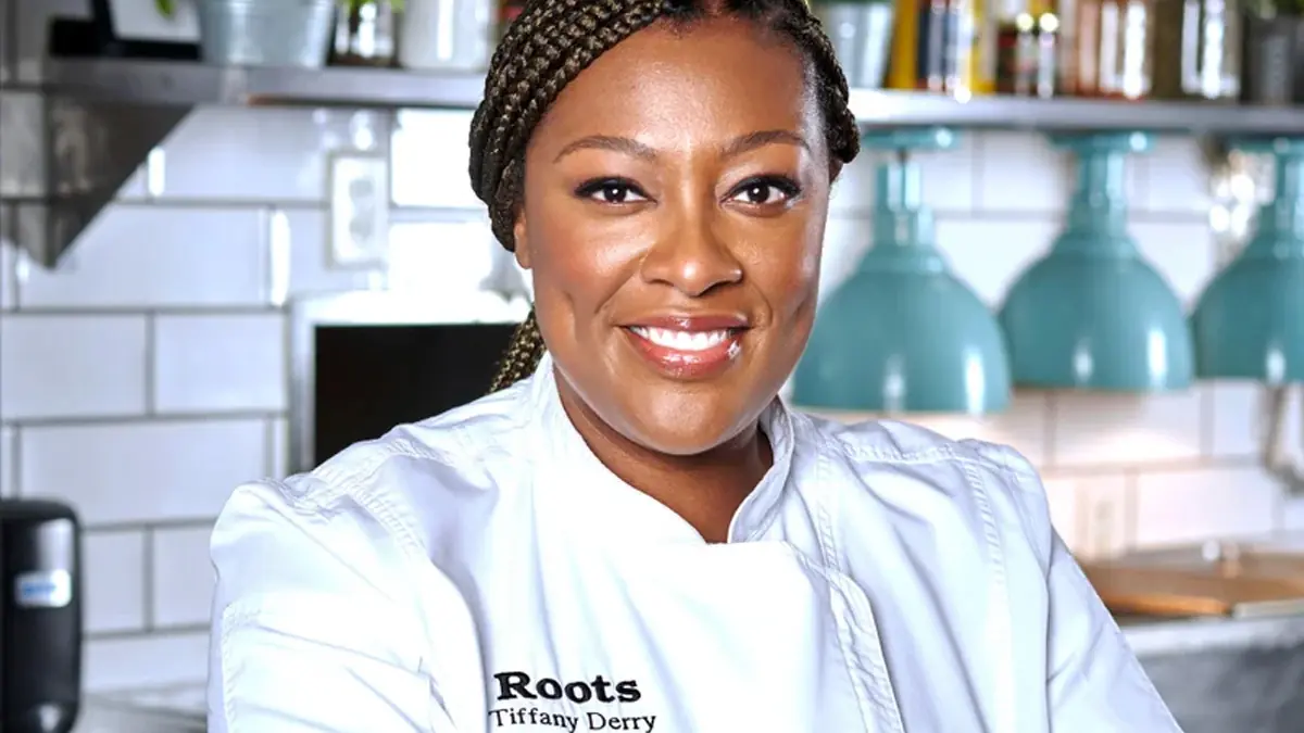 Chef in white Roots jacket stands in restaurant kitchen, background with teal lamps and shelves.