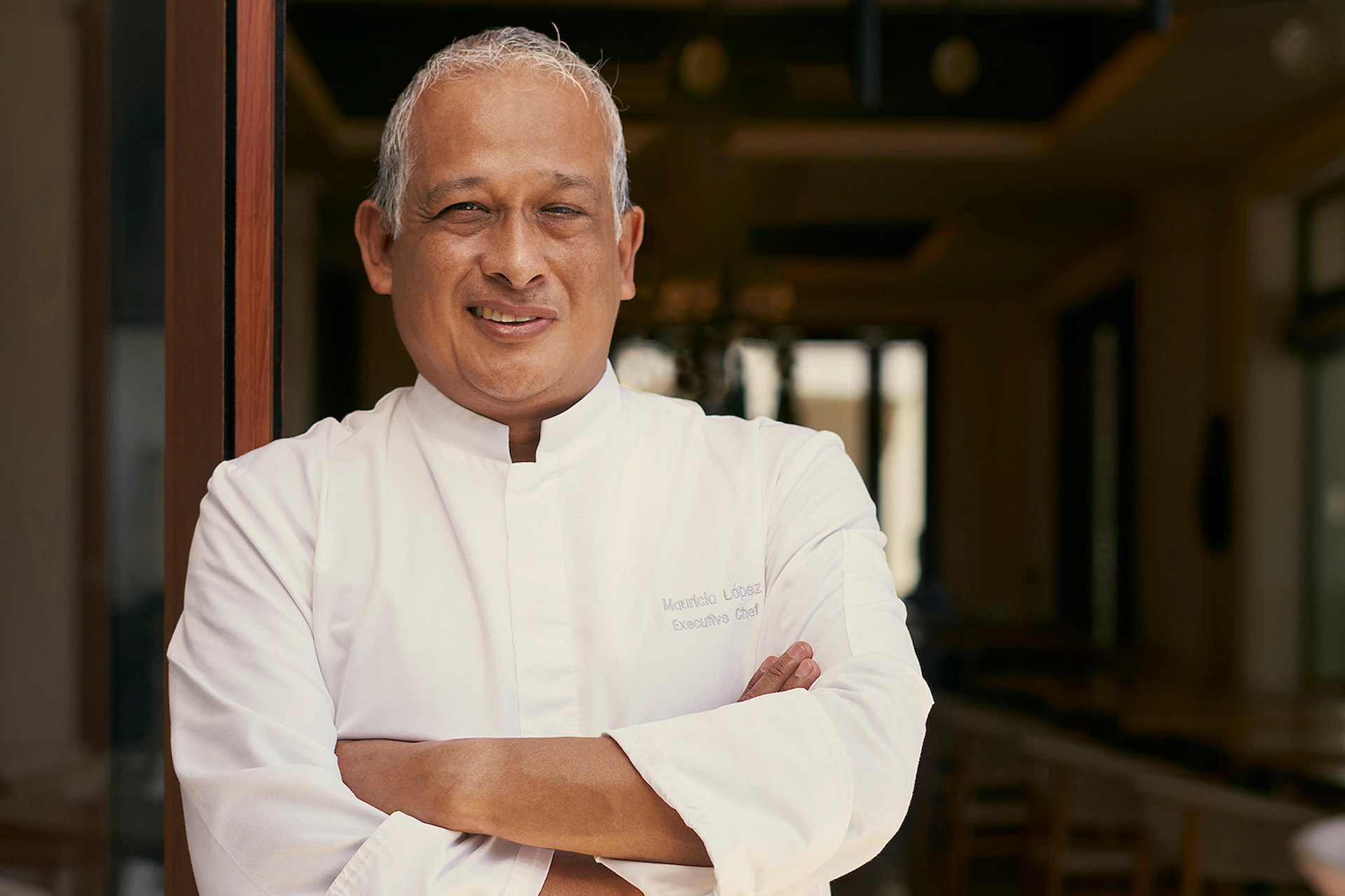 El chef Mauricio López, chef ejecutivo, de pie con confianza en el comedor de un restaurante, vistiendo una bata de chef blanca.