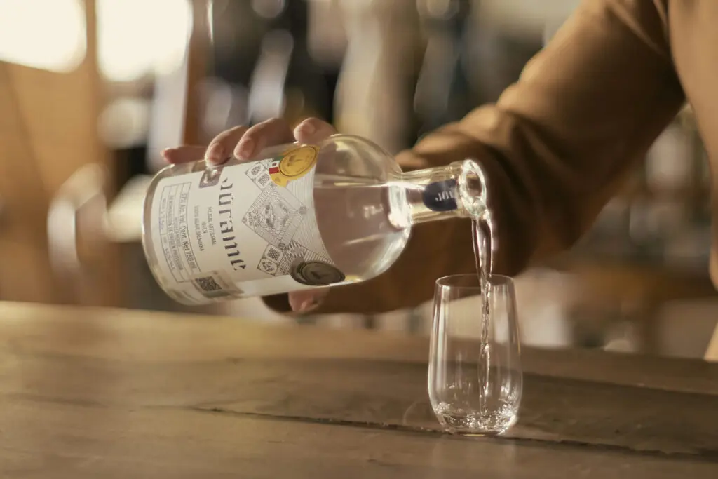 Hand pouring clear spirits from a glass bottle into a small tumbler on a wooden bar counter.
