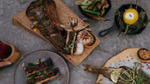 Presentación de comida gourmet con carnes a la parrilla, verduras y guarniciones servidas en platos y tablas rústicas.