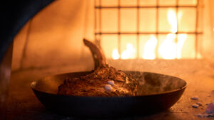 Filete Tomahawk chisporroteando en una sartén de hierro fundido dentro de un horno de leña con llamas brillando detrás.