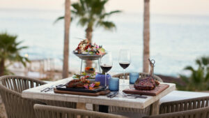 Elegante mesa de comedor al aire libre con torre de carnes y mariscos, copas de vino y vista al mar entre palmeras.