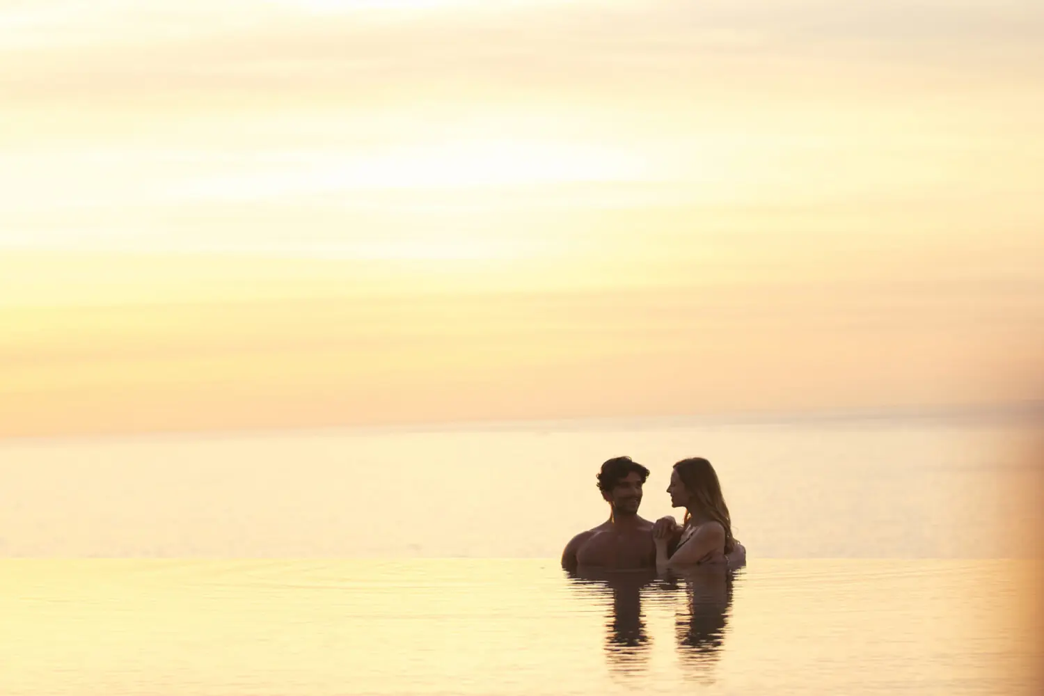 Una pareja silueteada disfruta de un momento romántico en una tranquila piscina infinita al atardecer con una suave luz dorada.