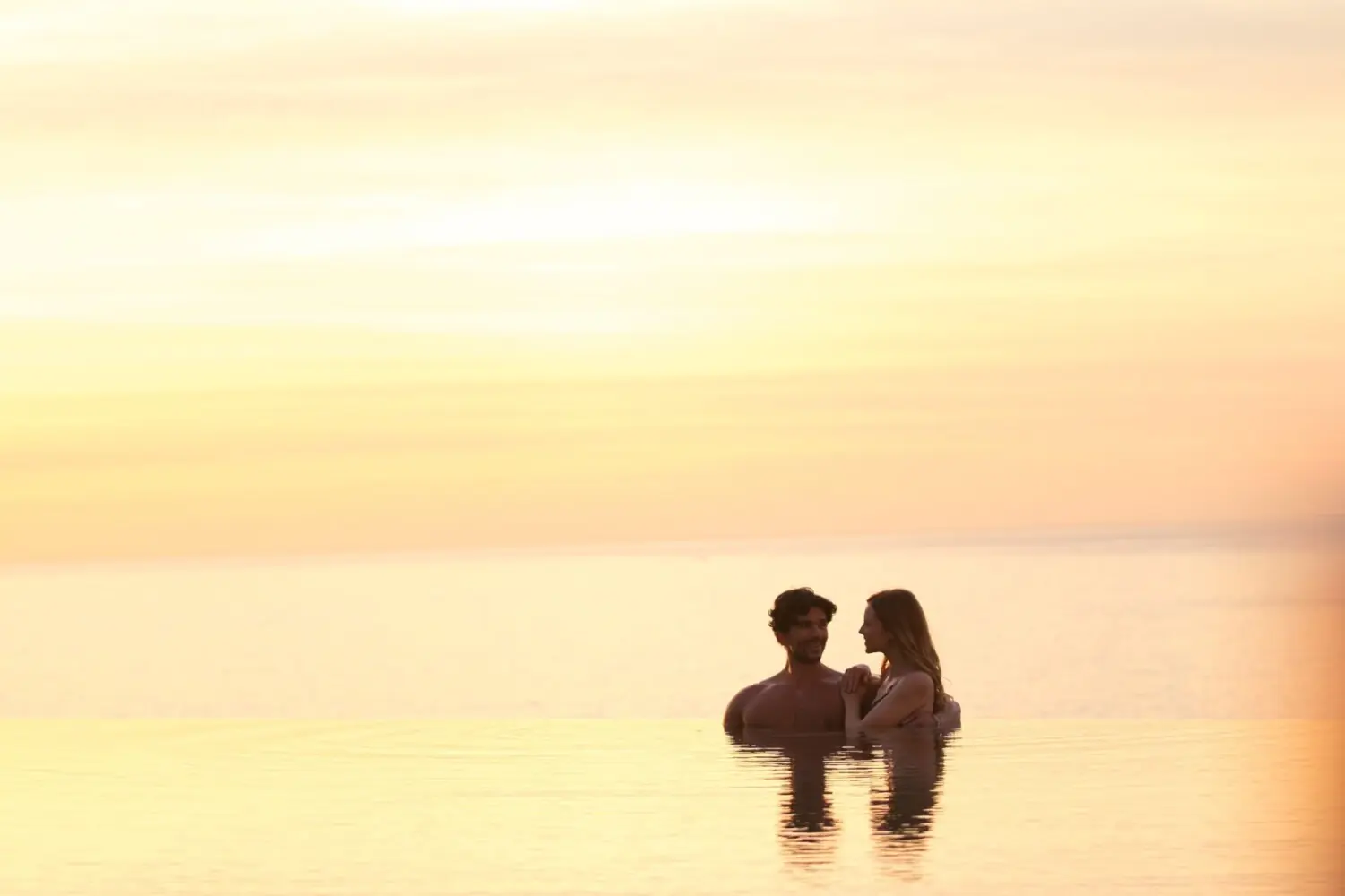Couple embraces in tranquil water, golden sunset sky reflecting on surface, romantic peaceful evening outdoors.