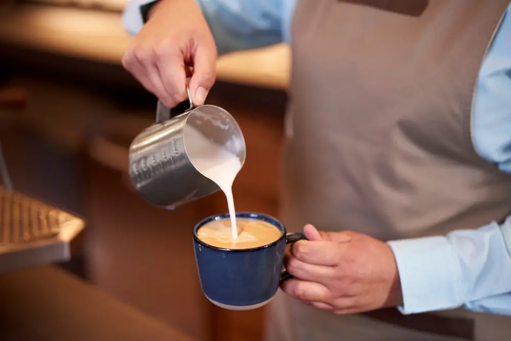 Barista pours steamed milk into coffee cup, creating latte art in cozy café setting.