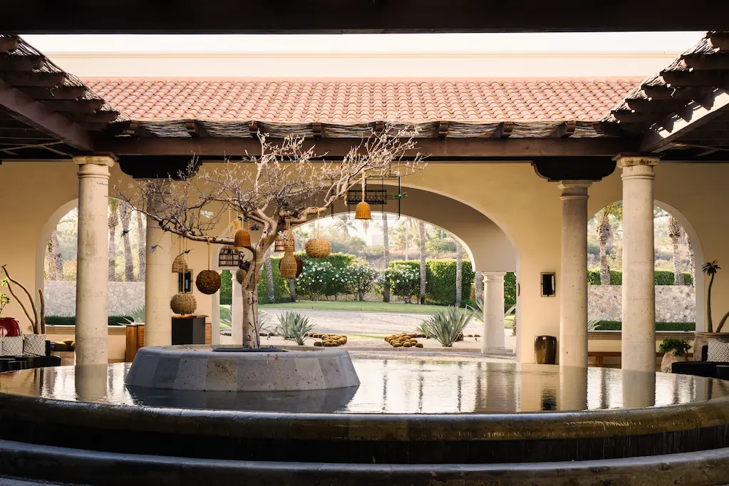 Open-air entryway with reflecting pool, decorated tree centerpiece, columns, arched walls, and views of lush gardens.
