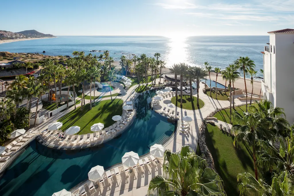 Aerial view of a beachfront resort with palm trees, winding pools, lounge chairs, and ocean views at sunrise.