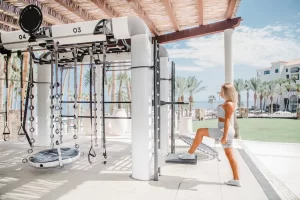 Mujer vestida con ropa deportiva utilizando equipos de gimnasio al aire libre, rodeada de palmeras y edificios del complejo bajo un cielo azul.