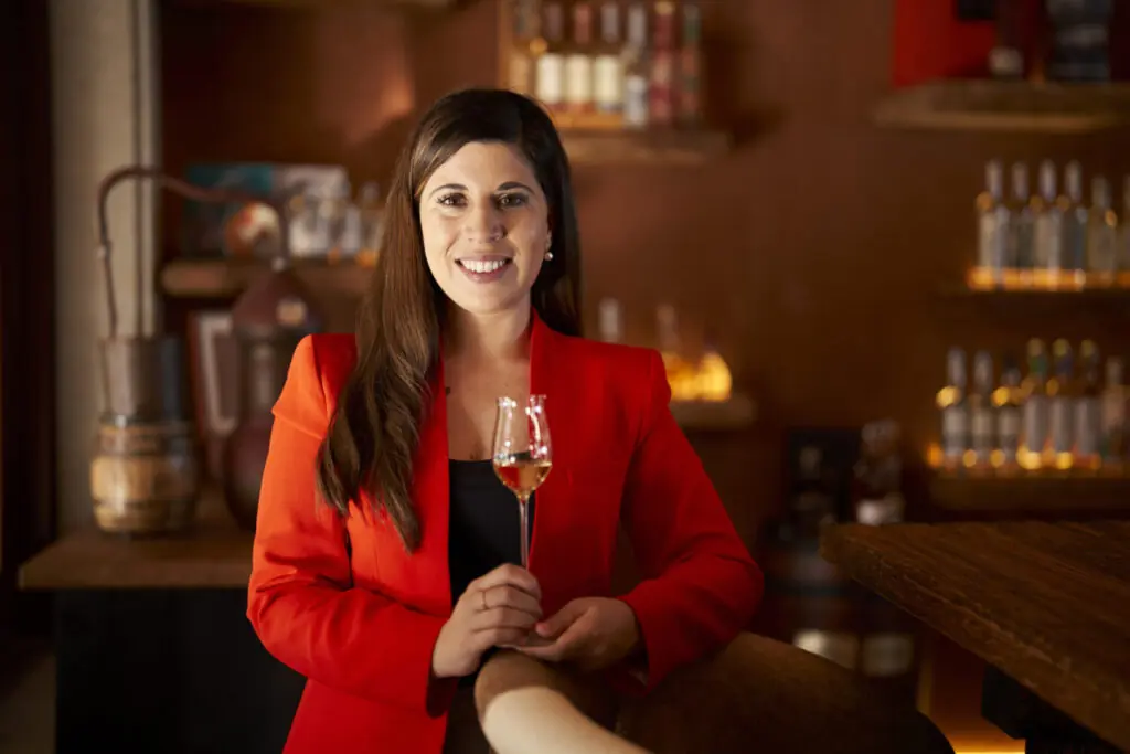 Woman in a red blazer holding a glass of liquor inside a warmly lit distillery or tasting room.