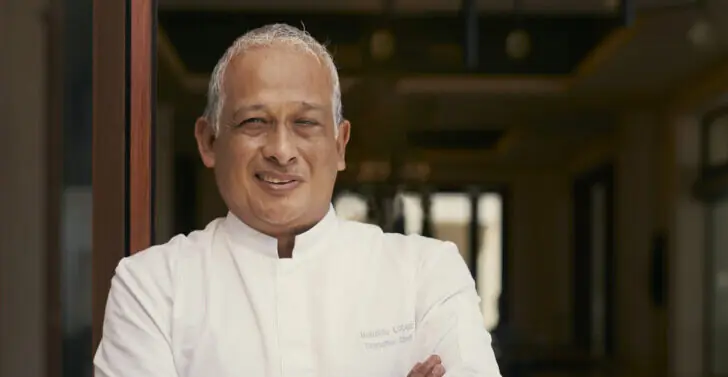 Smiling chef in a white uniform standing with arms crossed inside a restaurant.