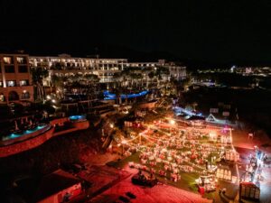 Large nighttime outdoor event at resort with string lights, dining tables, and illuminated pools, hotel buildings in background.