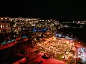 Gran evento nocturno al aire libre en el resort con luces de cadena, mesas de comedor y piscinas iluminadas, con edificios del hotel al fondo.