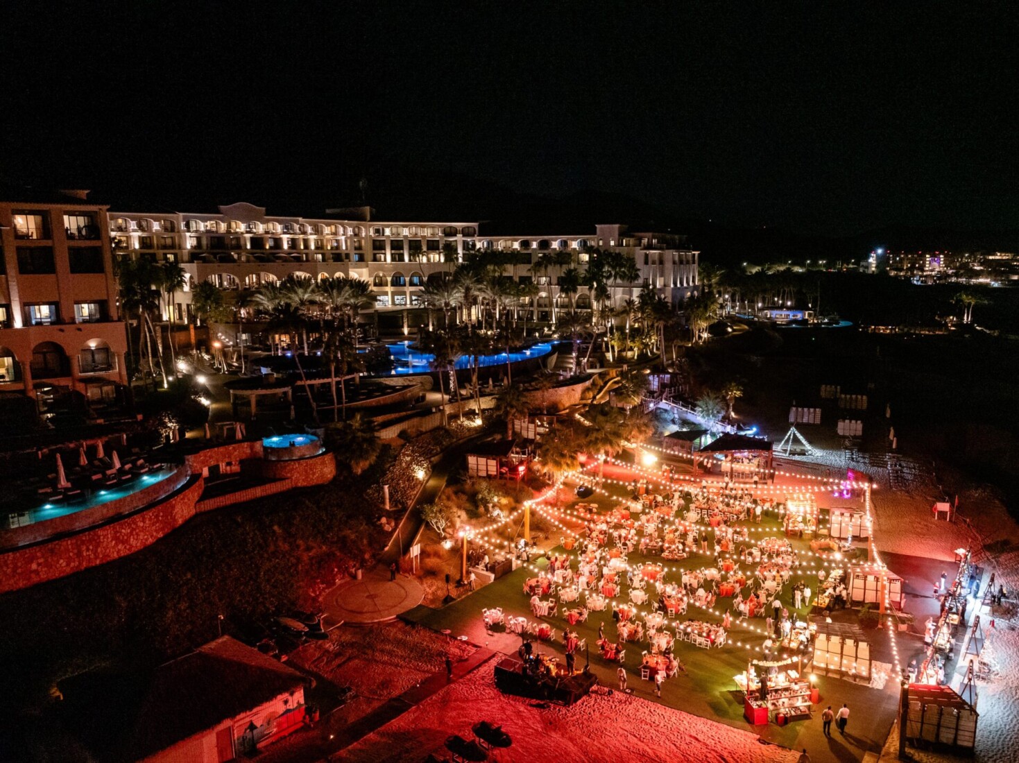 Large nighttime outdoor event at resort with string lights, dining tables, and illuminated pools, hotel buildings in background.