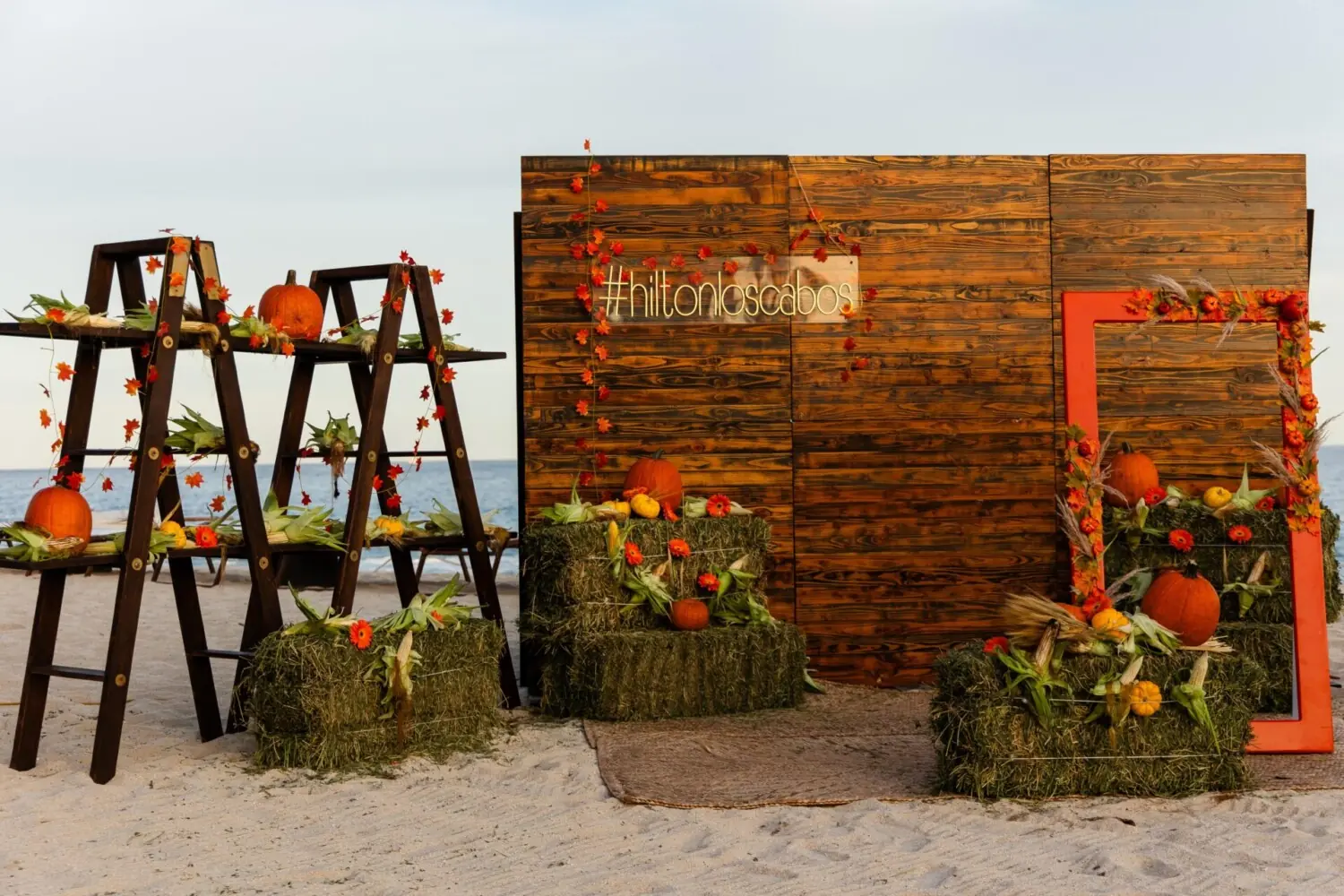 Exhibición rústica de playa con fondos de madera, calabazas, flores, maíz, fardos de heno y decoración de marcos de fotos de color naranja.