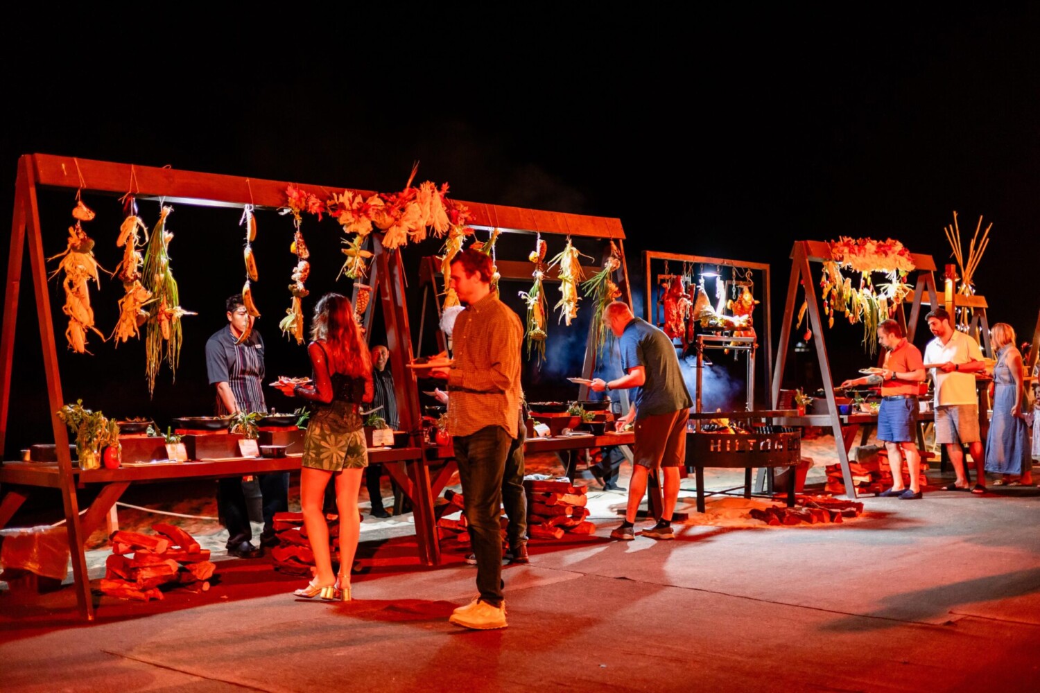 People selecting food at outdoor buffet stands decorated with hanging vegetables, under bright red lighting at night.
