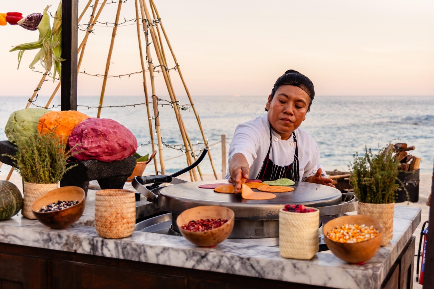 Chef prepares colorful handmade tortillas on outdoor grill, surrounded by fresh ingredients, near ocean backdrop.