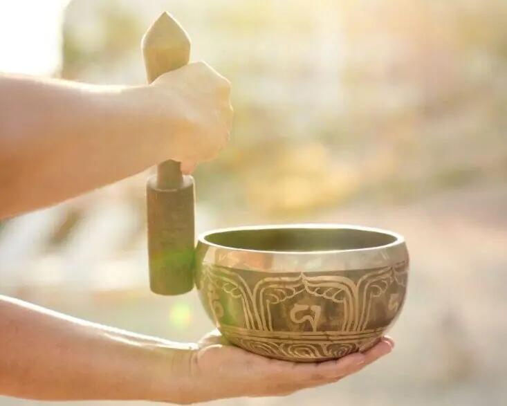 A person’s hands hold and play a decorative Tibetan singing bowl outside in warm, natural sunlight.