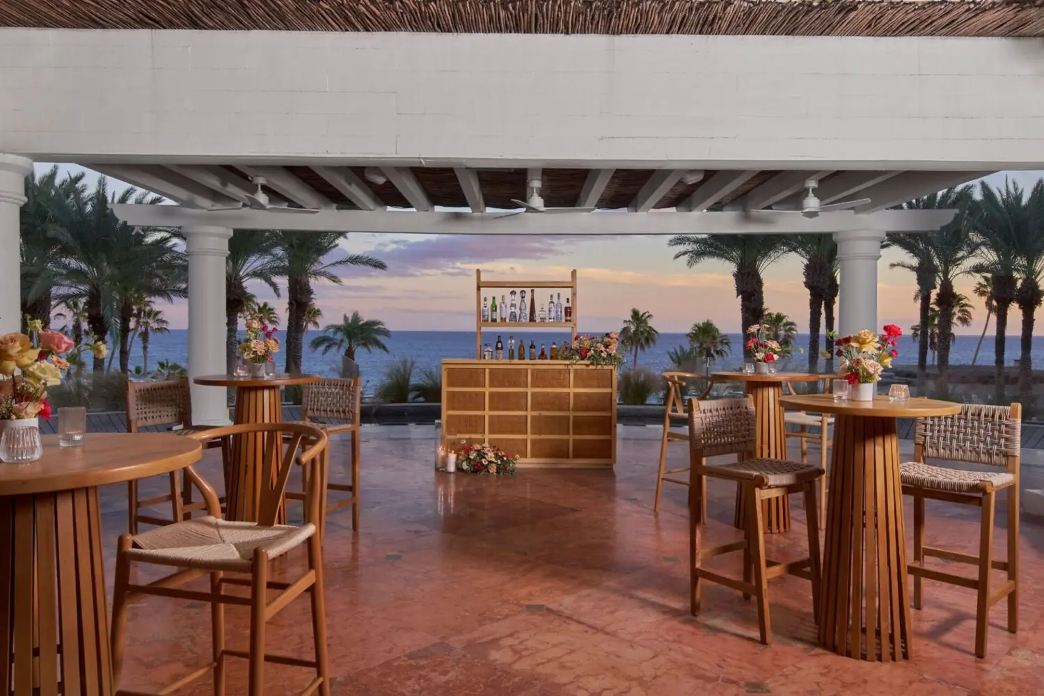 Elegant open-air bar with wooden tables and chairs, floral centerpieces, and ocean view at sunset, surrounded by palm trees.