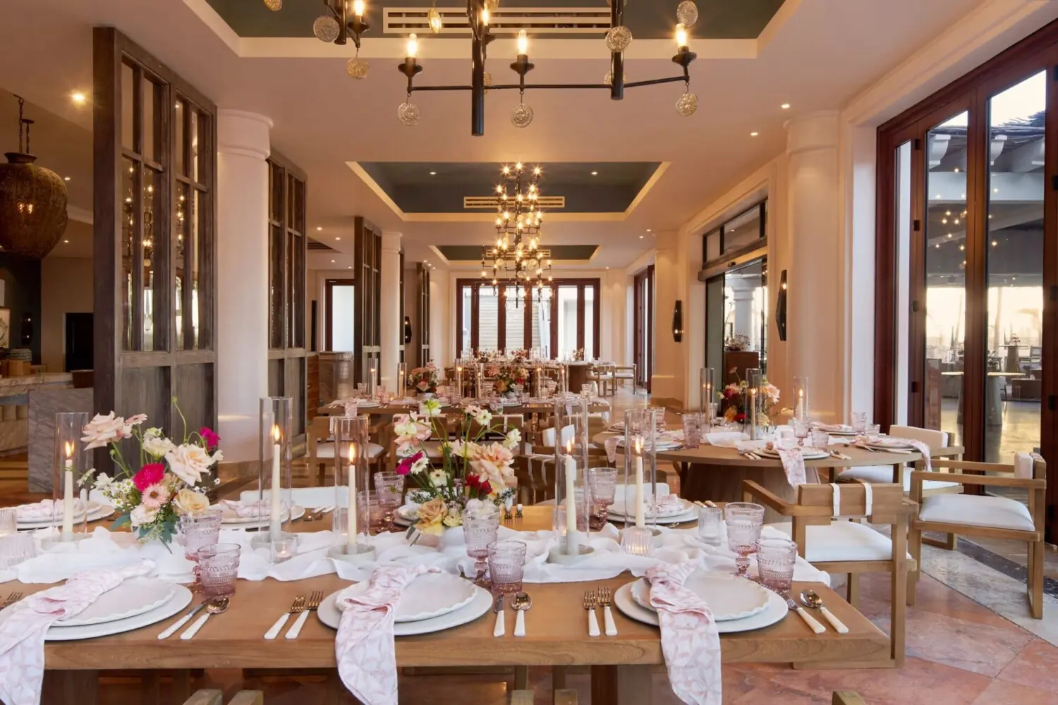 Elegant indoor wedding reception setup with wooden tables, flower centerpieces, candles, pink napkins, and natural light.