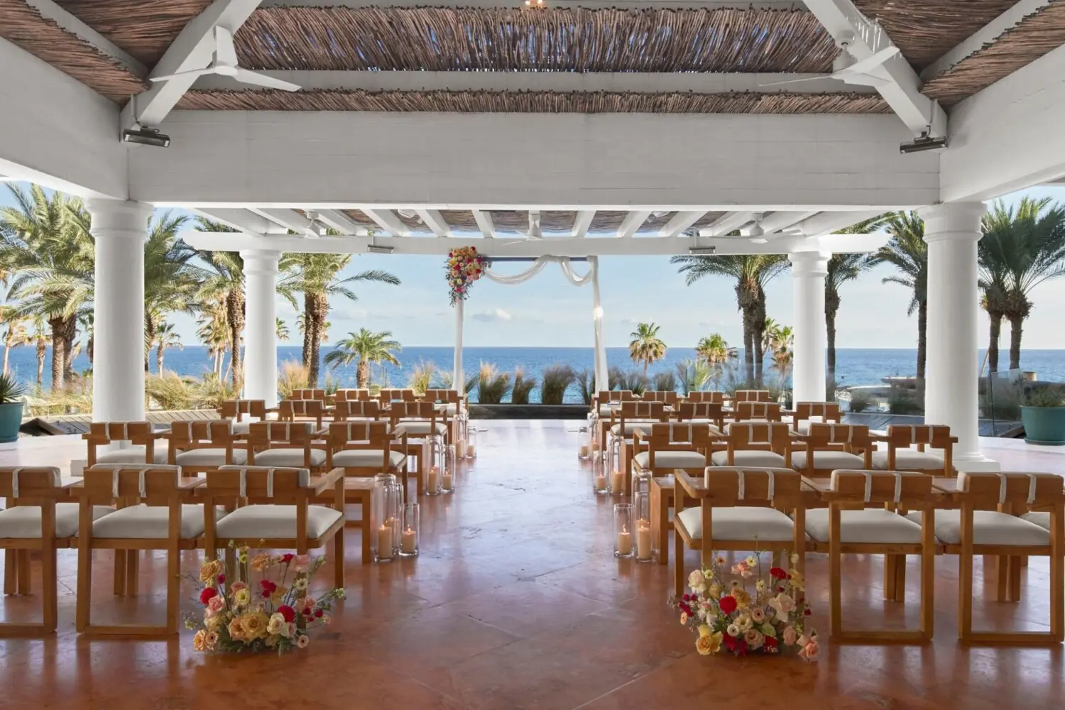 Indoor-outdoor wedding ceremony setup with wooden chairs, floral arrangements, sheer draped arbor, ocean and palm tree backdrop.