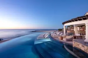 Infinity pool overlooking the ocean at sunset beside luxurious villa terrace with lounge chairs.
