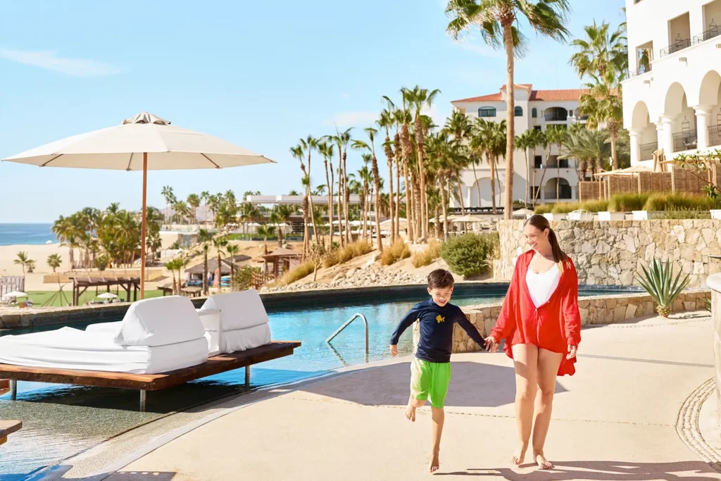 Mother and child walk hand-in-hand by resort poolside, palm trees and ocean in background.