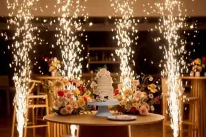 Wedding cake and floral arrangements on round table, indoor sparklers creating dazzling celebration effect behind.