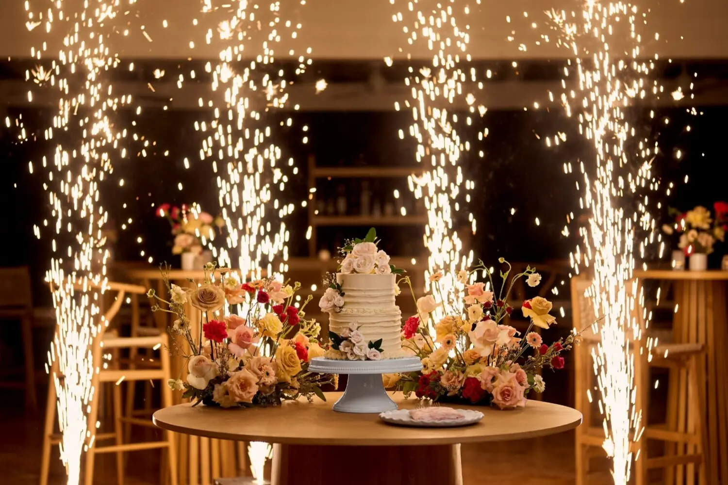 Wedding cake and floral arrangements on round table, indoor sparklers creating dazzling celebration effect behind.